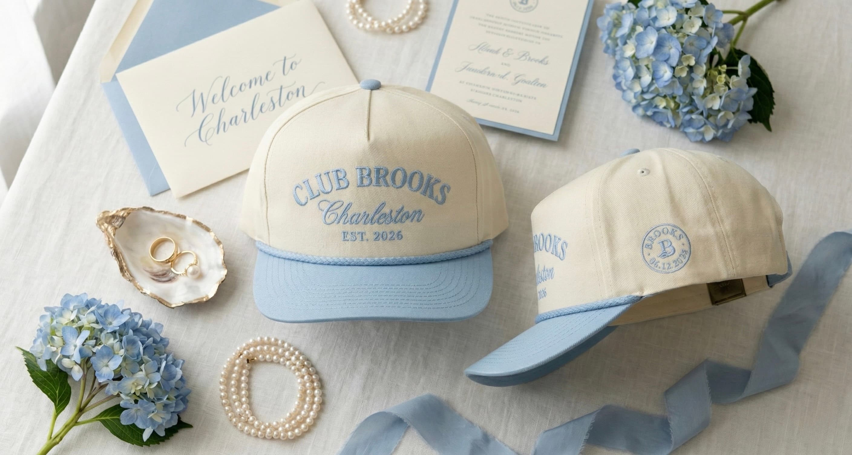 Cream rope caps with dusty-blue brims embroidered “Club Brooks, Charleston, Est. 2026”, beside a “Welcome to Charleston” card, an invitation, pearls and hydrangeas.