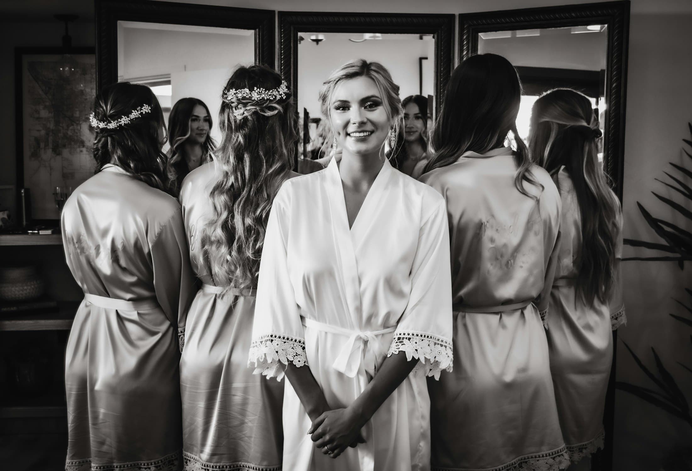 Black-and-white photograph of a bride in a white robe smiling, her bridesmaids in satin robes facing a row of mirrors behind her.